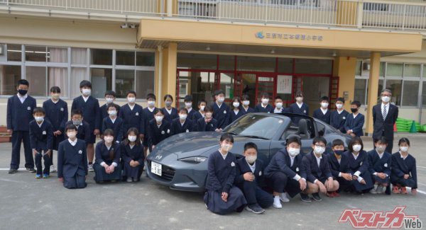 広島県三原市の本郷西小学校にて。山本氏と六年生の生徒たち。日本のスポーツカーを代表する４代目ロードスターとともに記念撮影