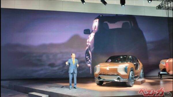 ジャパンモビリティショー2025三菱ブースでプレスブリーフィングに登壇する加藤隆雄CEO。写真右が世界初披露の「エレバンスコンセプト」。謎の新型車のシルエットが背景スクリーンに映し出された