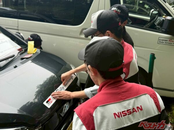 日産自動車大学校のメカニックさんたち。こちらも「若手育成枠」! 日産の作業ユニホームを着た若者が「GR」のステッカーをダイハツ ミライースDSRに貼るというモータースポーツらしい光景だ