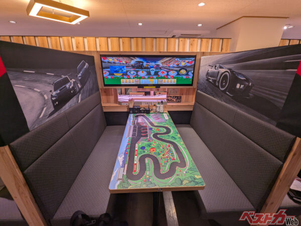 スシロー新宿西口店のテーブル席。机にサーキットが、大型ディスプレイにレースゲームが、左右の壁にGRマシンの写真が飾られる。クルマ好きならワクワクが止まらない空間