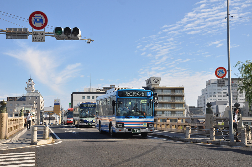 信号の左に一般車時間規制標識があり、続行するバスはスムーズに走を渡って駅方面へ