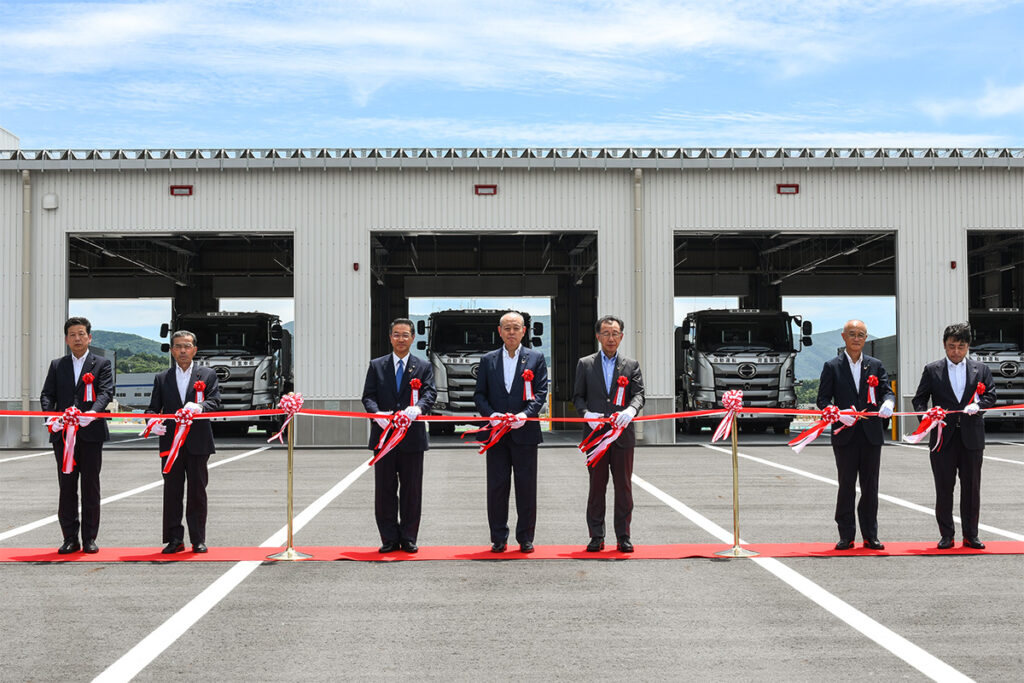 荷重車のガレージを背に記念式典を開催。大成ロテックの加賀田社長（右から2人目）に加えて、大成建設の谷山副社長（中央3人の中央）や田村市の白石市長（中央3人の向かって左）なども出席した。なお次世代技術実証センターには、大成建設の自然共生型管理棟や半自然草原の実証フィールドも設置予定
